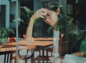 A young woman wearing glasses, sitting in a cafe with her hand on her head, looking pensive.