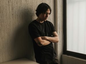Young man standing by a window with folded arms, looking thoughtful.