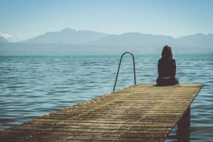 A person sitting alone on a wooden dock looking out at a large body of water and mountains.