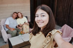 Smiling teenage girl taking a selfie with her parents sitting in the background, symbolizing family support during therapy decisions