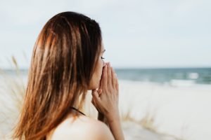 Woman with hands near face looking calm by the ocean, representing mindfulness and therapy for anxiety relief.