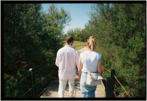 Back view of a couple walking hand in hand on a wooden bridge surrounded by greenery.